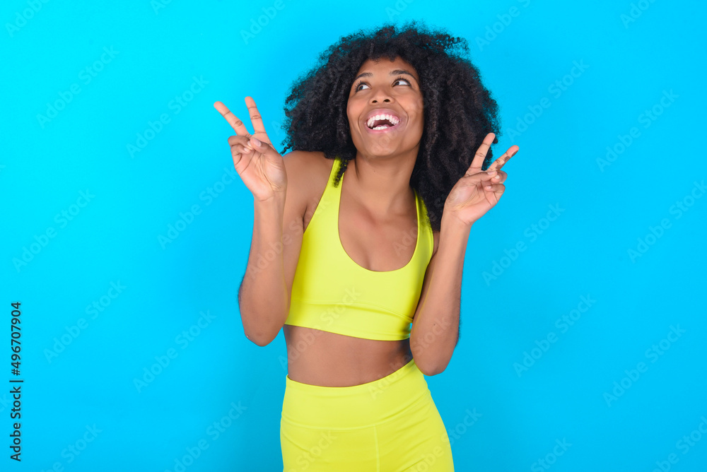 Isolated shot of cheerful young woman with afro hairstyle in sportswear against blue background makes peace or victory sign with both hands, feels cool. Isolated shot of cheerful young woman with afro hairstyle in sportswear against blue background makes peace or victory sign with both hands, feels cool.