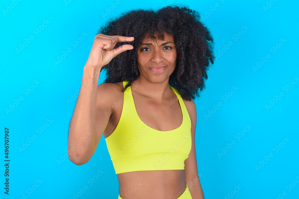 Upset young woman with afro hairstyle in sportswear against blue background shapes little gesture with hand demonstrates something very tiny small size. Not very much