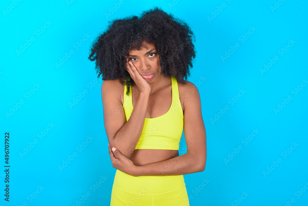Very bored young woman with afro hairstyle in sportswear against blue background holding hand on cheek while support it with another crossed hand, looking tired and sick. Very bored young woman with afro hairstyle in sportswear against blue background holding hand on cheek while support it with another crossed hand, looking tired and sick.