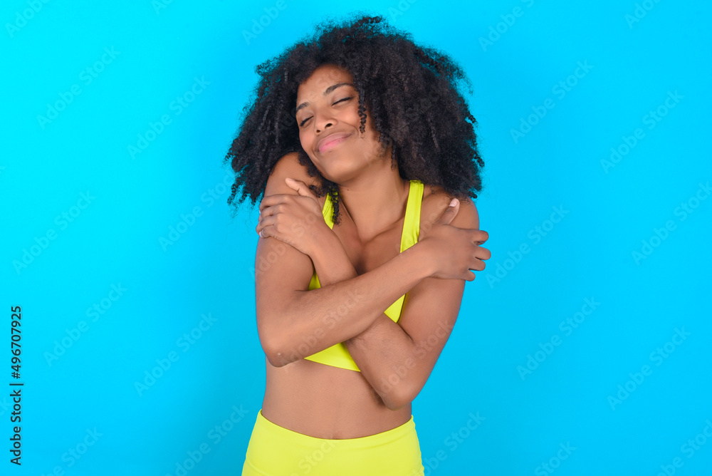 Naklejka premium young woman with afro hairstyle in sportswear against blue background Hugging oneself happy and positive, smiling confident. Self love and self care.