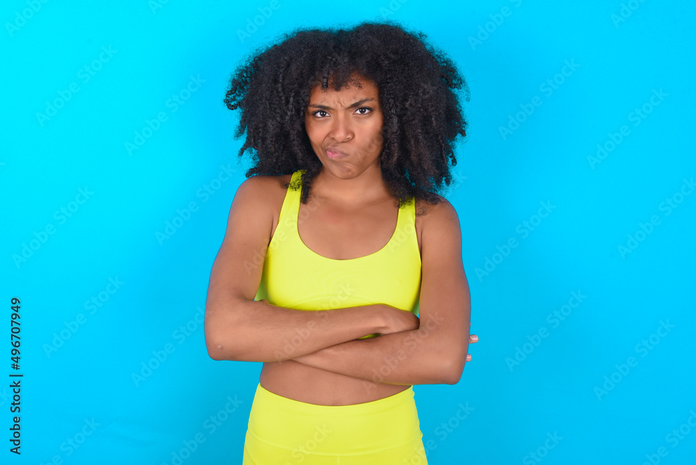 Picture of angry young woman with afro hairstyle in sportswear against blue background crossing arms. Looking at camera with disappointed expression. Picture of angry young woman with afro hairstyle in sportswear against blue background crossing arms. Looking at camera with disappointed expression.