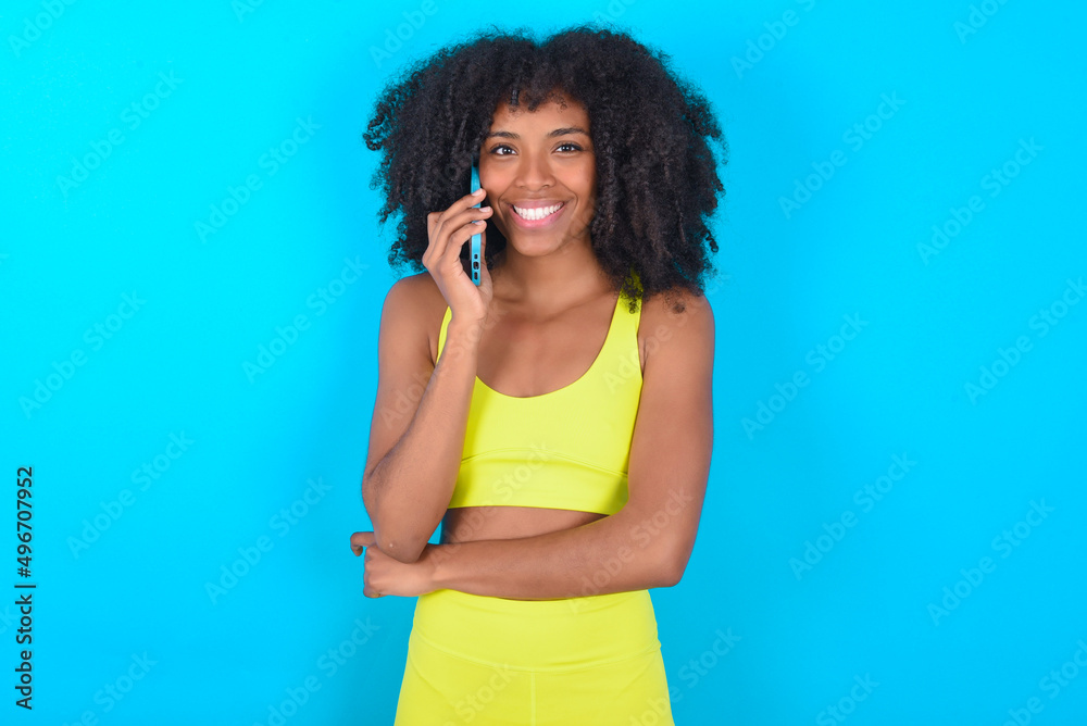 Portrait of a smiling young woman with afro hairstyle in sportswear against blue background talking on mobile phone. Business, confidence and communication concept. Portrait of a smiling young woman with afro hairstyle in sportswear against blue background talking on mobile phone. Business, confidence and communication concept.