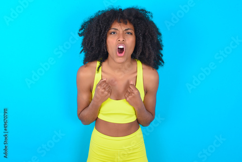 Joyful excited lucky young woman with afro hairstyle in sportswear against blue background cheering, celebrating success, screaming yes with clenched fists