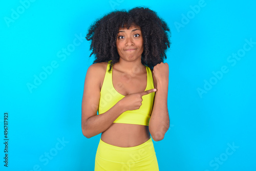 young woman with afro hairstyle in sportswear against blue background in hurry pointing to watch time, impatience, upset and angry for deadline delay