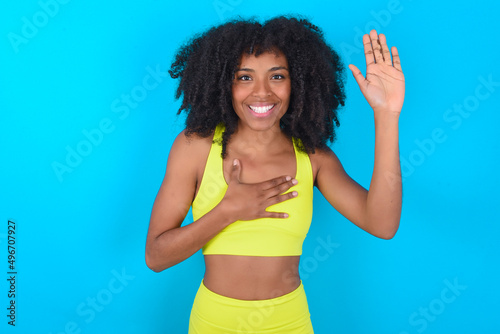 I swear, promise you not regret. Portrait of sincere young woman with afro hairstyle in sportswear against blue raising one arm and hold hand on heart as give oath, telling truth, want you to believe.