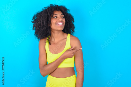 Happy cheerful smiling young woman with afro hairstyle in sportswear against blue background looking and pointing aside with hand. Copy space and advertisement concept.