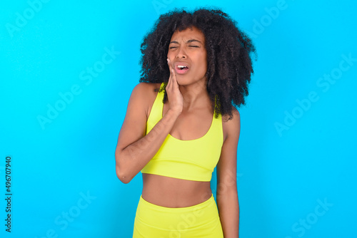 Tooth ache concept. young woman with afro hairstyle in sportswear against blue wall feeling pain, holding his cheek with hand, suffering from bad toothache, looking at camera with painful expression