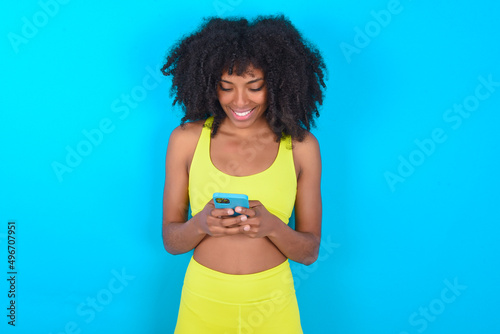 Smiling young woman with afro hairstyle in sportswear against blue wall using cell phone, messaging, being happy to text with friends, looking at smartphone. Modern technologies and communication.