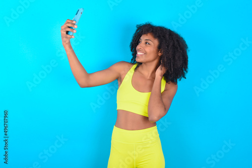 Portrait of a young woman with afro hairstyle in sportswear against blue background taking a selfie to send it to friends and followers or post it on his social media.