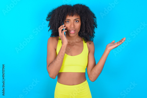 young woman with afro hairstyle in sportswear against blue wall talking on the phone stressed with hand on face, shocked with shame and surprise face, angry and frustrated. Fear and upset for mistake.