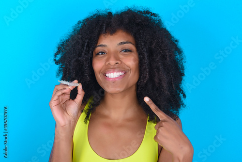 young woman with afro hairstyle in sportswear against blue background holding an invisible aligner and pointing to her perfect straight teeth. Dental healthcare and confidence concept. photo