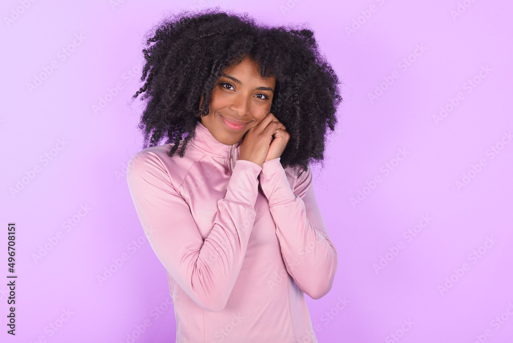Charming serious young woman with afro hairstyle in technical sports shirt against purple background keeps hands near face smiles tenderly at camera Charming serious young woman with afro hairstyle in technical sports shirt against purple background keeps hands near face smiles tenderly at camera