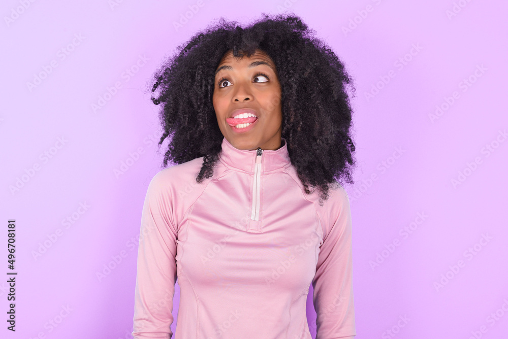 Funny young woman with afro hairstyle in technical sports shirt against purple background makes grimace and crosses eyes plays fool has fun alone sticks out tongue. Funny young woman with afro hairstyle in technical sports shirt against purple background makes grimace and crosses eyes plays fool has fun alone sticks out tongue.