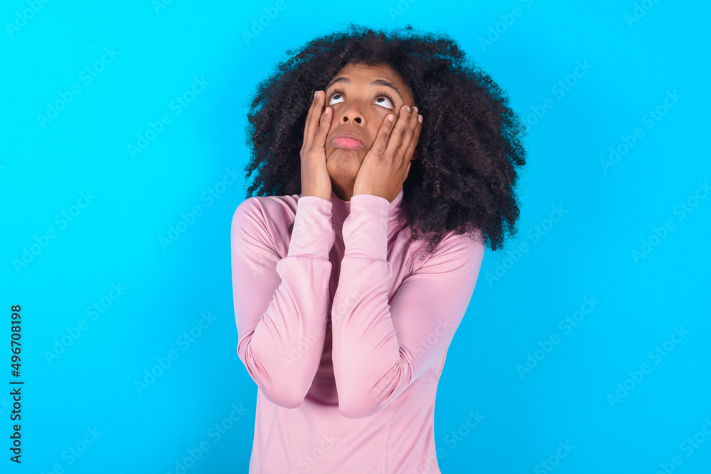 young woman with afro hairstyle in technical sports shirt against blue background keeps hands on cheeks has bored displeased expression. Stressed hopeless model young woman with afro hairstyle in technical sports shirt against blue background keeps hands on cheeks has bored displeased expression. Stressed hopeless model