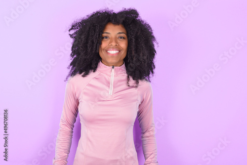 young woman with afro hairstyle in technical sports shirt against purple background with nice beaming smile pleased expression. Positive emotions concept