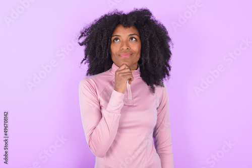 Thoughtful young woman with afro hairstyle in technical sports shirt against purple background holds chin and looks away pensively makes up great plan