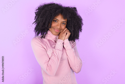 Charming serious young woman with afro hairstyle in technical sports shirt against purple background keeps hands near face smiles tenderly at camera