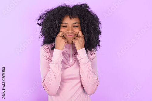 Pleased young woman with afro hairstyle in technical sports shirt against purple background with closed eyes keeps hands near cheeks and smiles tenderly imagines something very pleasant