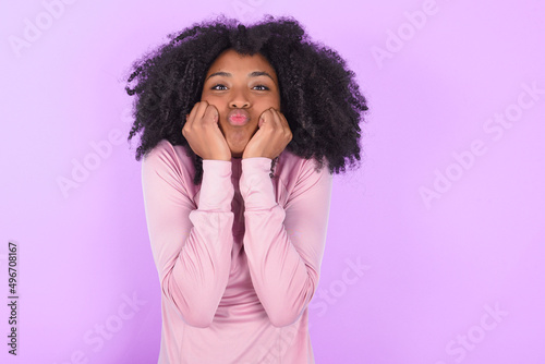 young woman with afro hairstyle in technical sports shirt against purple background with surprised expression keeps hands under chin keeps lips folded makes funny grimace