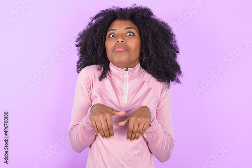 young woman with afro hairstyle in technical sports shirt against purple background makes bunny paws and looks with innocent expression plays with her little kid