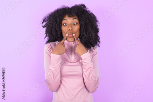 young woman with afro hairstyle in technical sports shirt against purple background crosses eyes and makes fish lips funny grimace