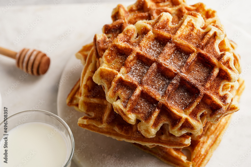 Sweet breakfast - fresh homemade belgian waffles with honey and powdered sugar stacked on white round marble board and small glass with milk Sweet breakfast - fresh homemade belgian waffles with honey and powdered sugar stacked on white round marble board and small glass with milk