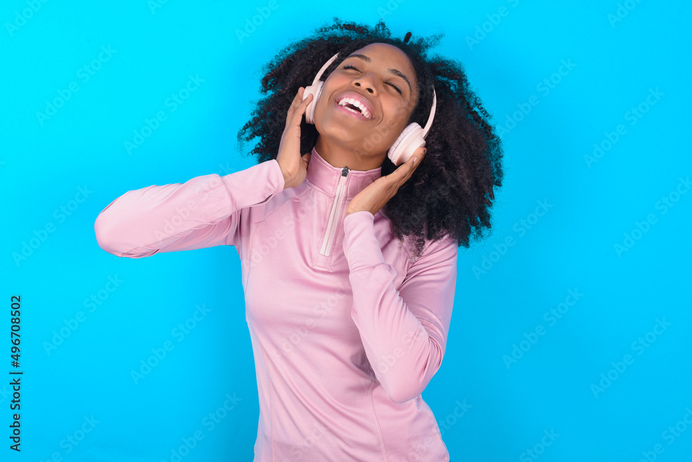 young woman with afro hairstyle in technical sports shirt against blue background smiles broadly feels very glad listens favourite music track via wireless headphones closes eyes. young woman with afro hairstyle in technical sports shirt against blue background smiles broadly feels very glad listens favourite music track via wireless headphones closes eyes.