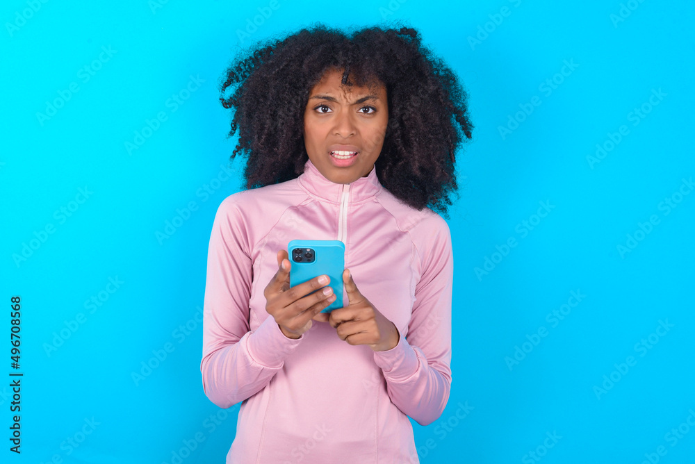 Photo of astonished crazy young woman with afro hairstyle in technical sports shirt against blue background hold smartphone dislike feedback concept Photo of astonished crazy young woman with afro hairstyle in technical sports shirt against blue background hold smartphone dislike feedback concept