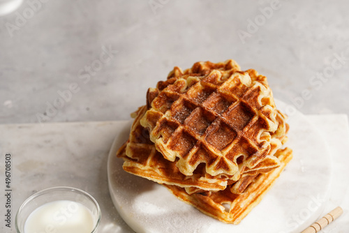 Sweet breakfast - fresh homemade belgian waffles with honey and powdered sugar stacked on white round marble board and small glass with milk photo