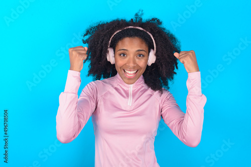 Emotional young woman with afro hairstyle in technical sports shirt against blue background exclaims loudly feels like winner raises clenched fists keeps mouth opened wears stereo headphones on ears 