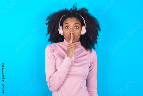 young woman with afro hairstyle in technical sports shirt against blue background making hush gesture with finger on her lips wearing wireless headphones. Be quiet.