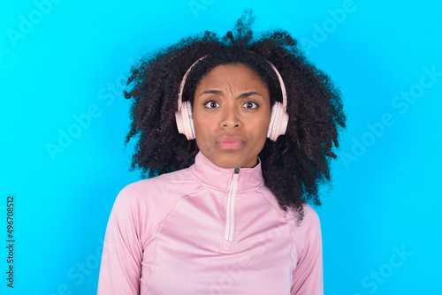 Serious displeased young woman with afro hairstyle in technical sports shirt against blue background looks puzzled at camera being angry wears stereo headphones listens music while walking at street © Jihan