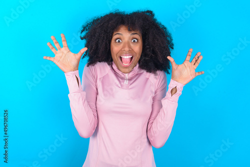 Delighted positive young woman with afro hairstyle in technical sports shirt against blue background opens mouth and arms palms up after having great result
