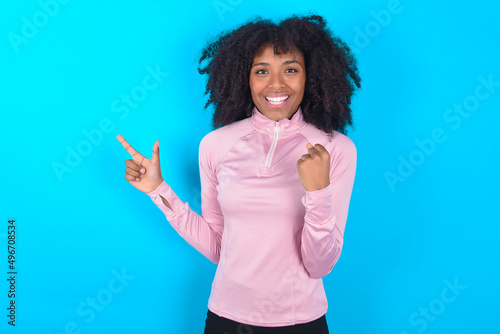 young woman with afro hairstyle in technical sports shirt against blue background points at empty space holding fist up, winner gesture.
