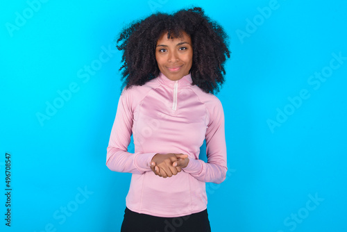 Photo of cheerful confident young woman with afro hairstyle in technical sports shirt against blue background arms together