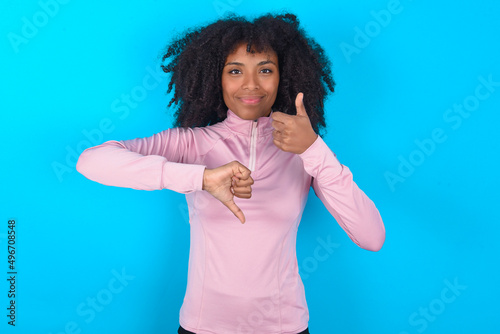young woman with afro hairstyle in technical sports shirt against blue background showing thumb up down sign