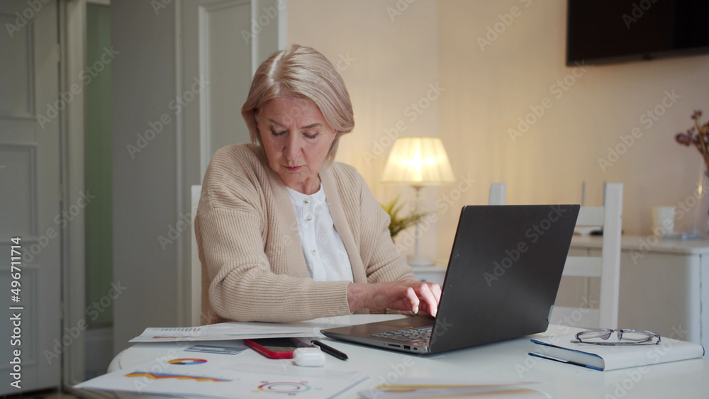 An old lady is doing paperwork on a laptop. She studies diagrams and blueprints An old lady is doing paperwork on a laptop. She studies diagrams and blueprints