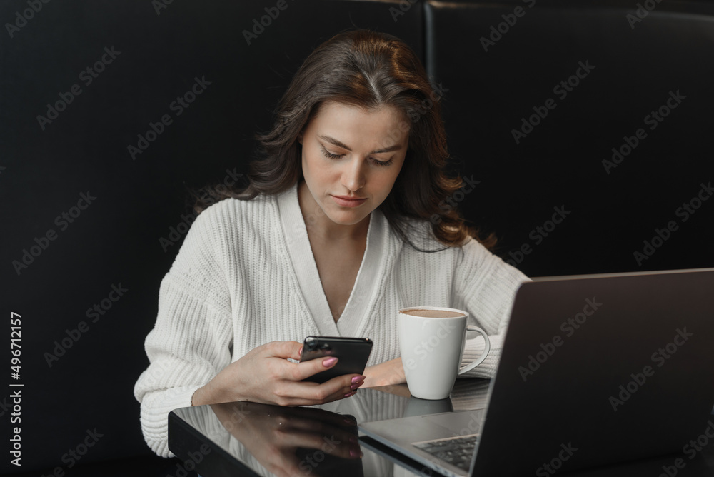 A young concentrated woman in a cafe reads a text message or news from her mobile phone, sits at a table in a cafe with a laptop. A young concentrated woman in a cafe reads a text message or news from her mobile phone, sits at a table in a cafe with a laptop.
