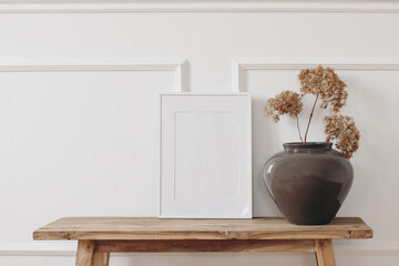 Brown bold vase. Dry hydrangea flowers on old wooden bench. Blank black picture frame mockup. White wall moulding background, stucco decor. Empty copy space. Elegant interior. Summer, fall still life