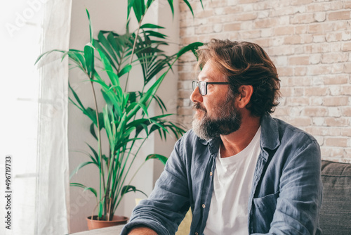 Thoughtful mature man looking out through window sitting on sofa at modern luxury apartment. Contemplative caucasian male admiring view sitting comfortably on couch at home