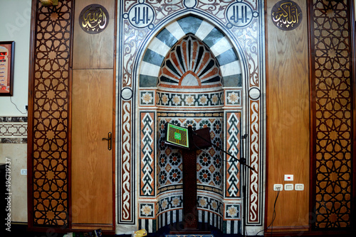 A mosque from inside with Islamic decorative style, mosque Mihrab or niche with a Quran put on a wooden stand and a microphone, translation of Arabic text (Allah, God is the greatest), selective focus photo