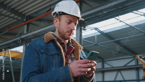 Close up handsome engineer man with helmet and uniform use mobile phone standing in factory. Communication, worker. photo