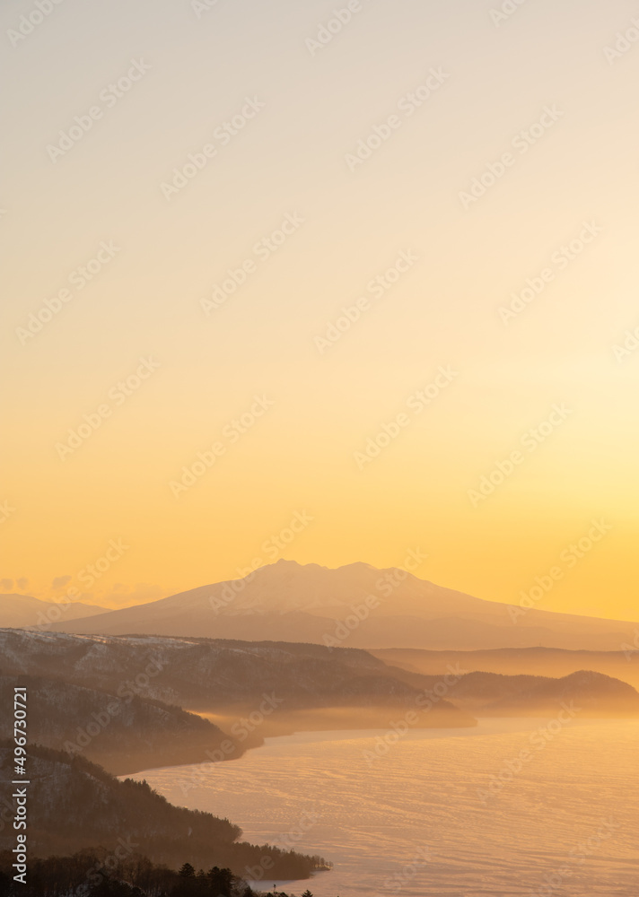 薄いオレンジ色の夜明けの空の下の霞んだ山々のシルエットと湖の風景。