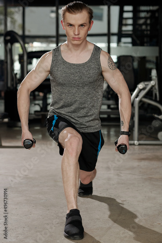 Strong young man holding dumbbells in both hands when doing forward lunges in gym