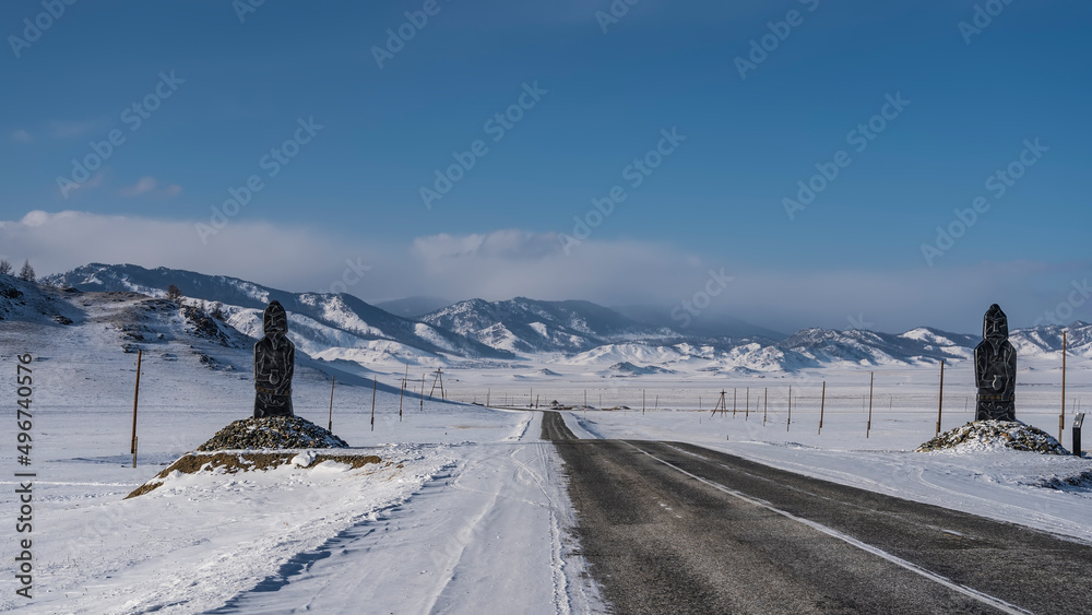 The highway goes off into the distance through a snow-covered valley. White markings on the asphalt. The figures of idols stand on the roadsides. Ahead, against the sky-picturesque mountains. Altai. The highway goes off into the distance through a snow-covered valley. White markings on the asphalt. The figures of idols stand on the roadsides. Ahead, against the sky-picturesque mountains. Altai.