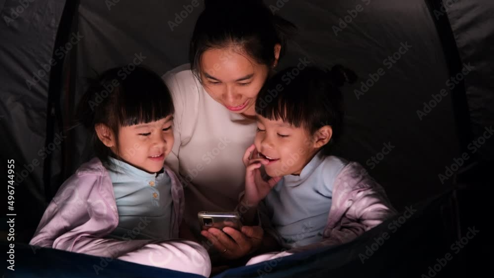 Young mother and her children play on smartphones while sitting in a tent at night. Funny mom and lovely child having fun with mobile phones. Happy family spent together on vacation. Young mother and her children play on smartphones while sitting in a tent at night. Funny mom and lovely child having fun with mobile phones. Happy family spent together on vacation.