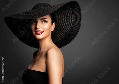 Cheerful Smiling Woman in Summer Hat. Beauty Model in Big Black Hat and Dress. Fashion Girl Face Portrait with Red Lipstick. Elegant Lady over dark Gray Background © inarik