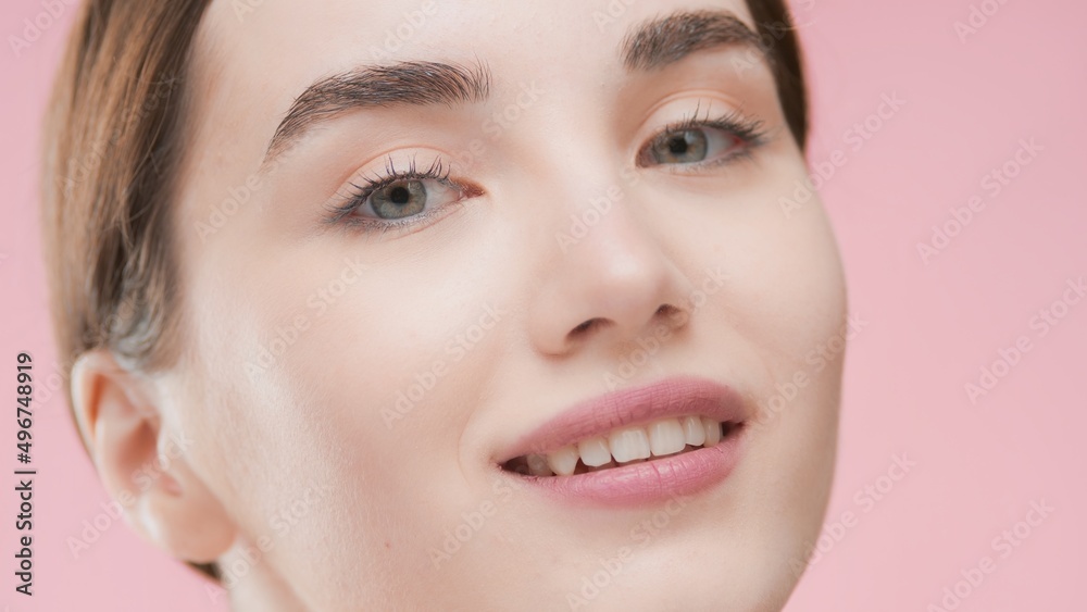 Extreme close-up beauty portrait of young fit attractive Caucasian woman with brown who smiles and looks at camera against pink background | Skin care concept
