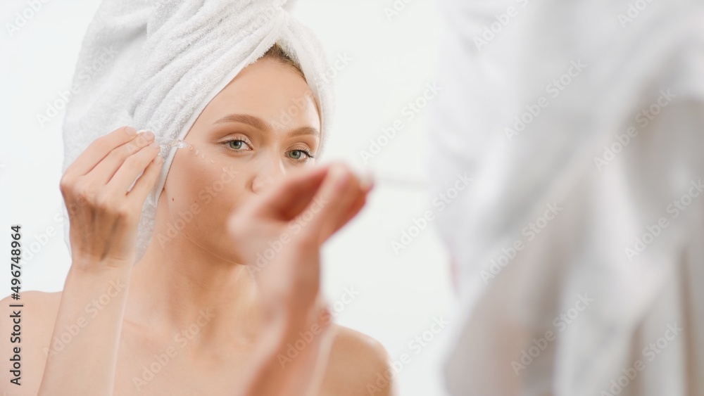 Young woman wrapped in a towel applies skin care serum on her face using a dropper in front of the mirror in the bathroom | Face serum commercial concept Young woman wrapped in a towel applies skin care serum on her face using a dropper in front of the mirror in the bathroom | Face serum commercial concept