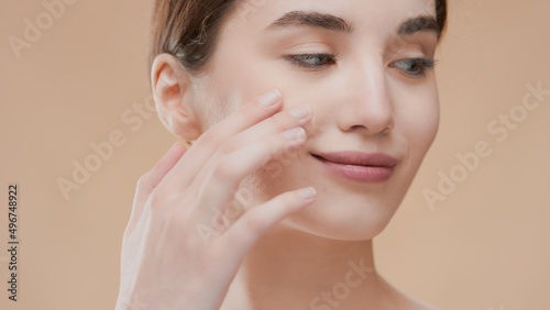 Attractive young brunette Caucasian woman gently touches her face with fingers looking aside on beige ripple background | Face care product commercial